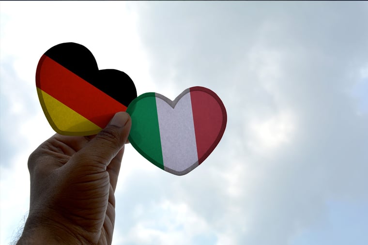Hand holding two paper hearts, one with the German flag and the other with the Italian flag